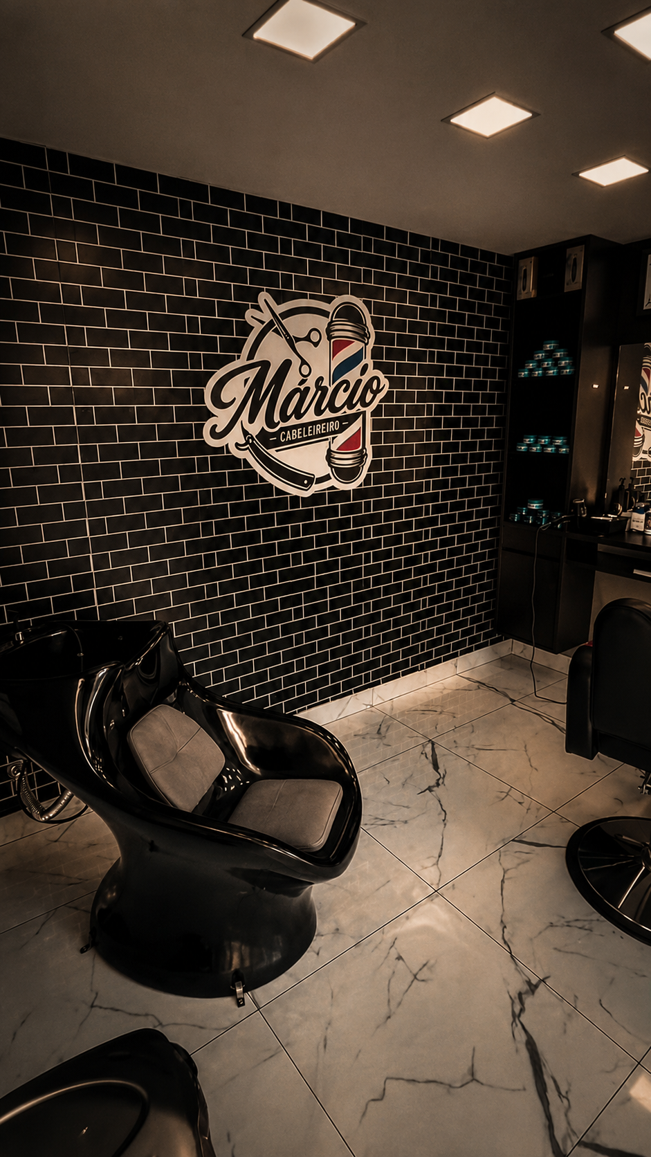 Interior do salão Márcio Cabeleireiro: parede de tijolinho preto com o logo iluminado, piso de mármore branco, cadeira de lavagem e prateleira de produtos.